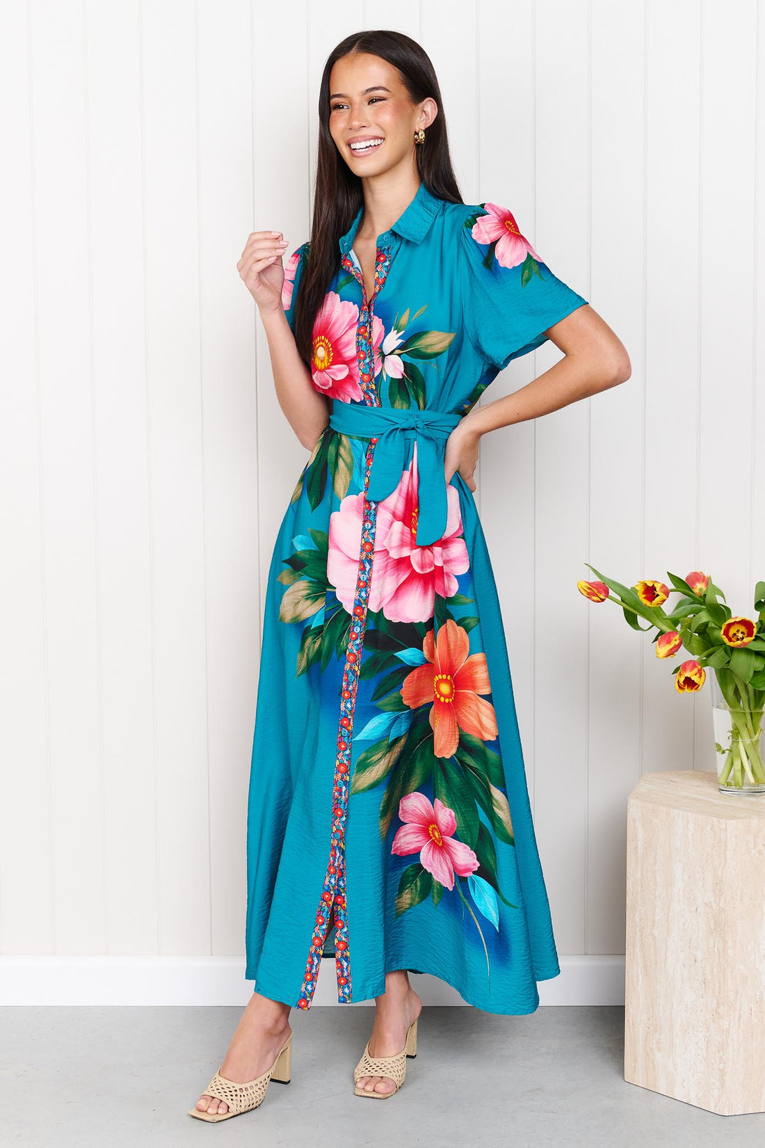 A woman wearing the minnie + MAXXIE Mona Maxi Dress in bright teal blue with a large floral print and waist tie, standing in front of a white panelled wall.