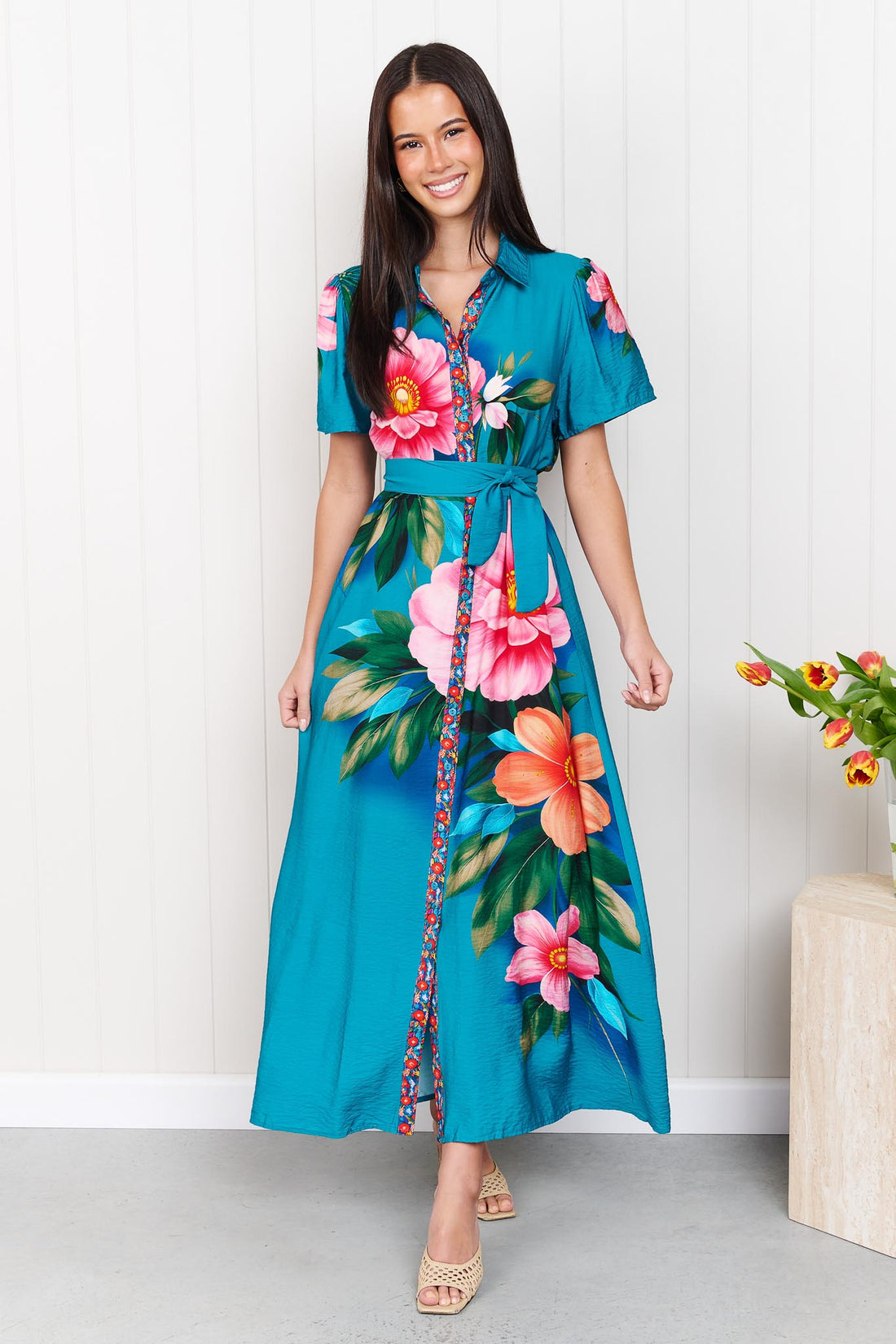 A woman wearing the minnie + MAXXIE Mona Maxi Dress in bright teal blue with a large floral print and waist tie, standing in front of a white panelled wall.