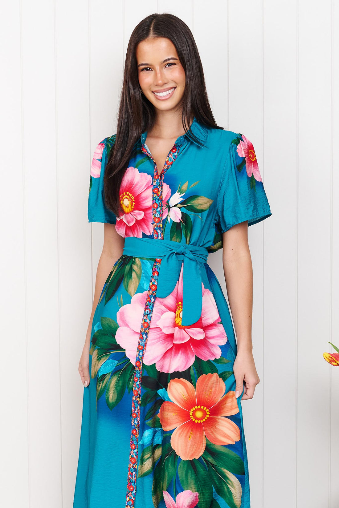 A woman wearing the minnie + MAXXIE Mona Maxi Dress in bright teal blue with a large floral print and waist tie, standing in front of a white panelled wall.