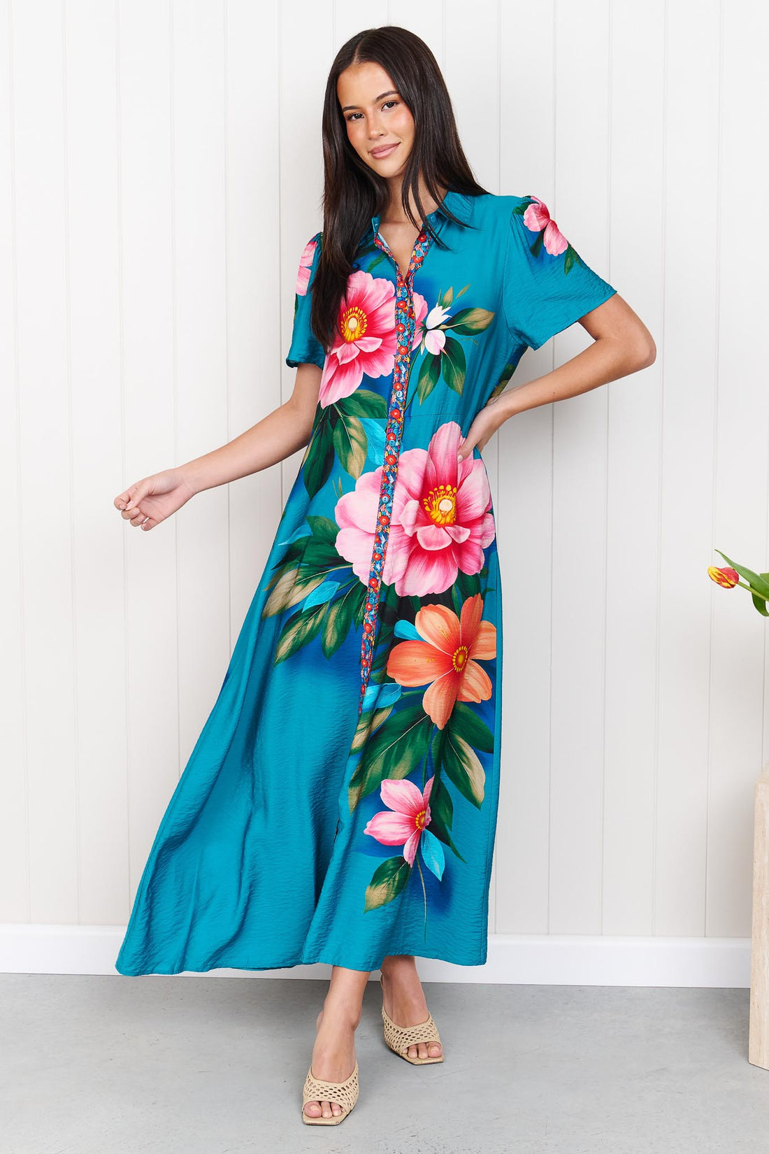A woman wearing the minnie + MAXXIE Mona Maxi Dress in bright teal blue with a large floral print, standing in front of a white panelled wall.