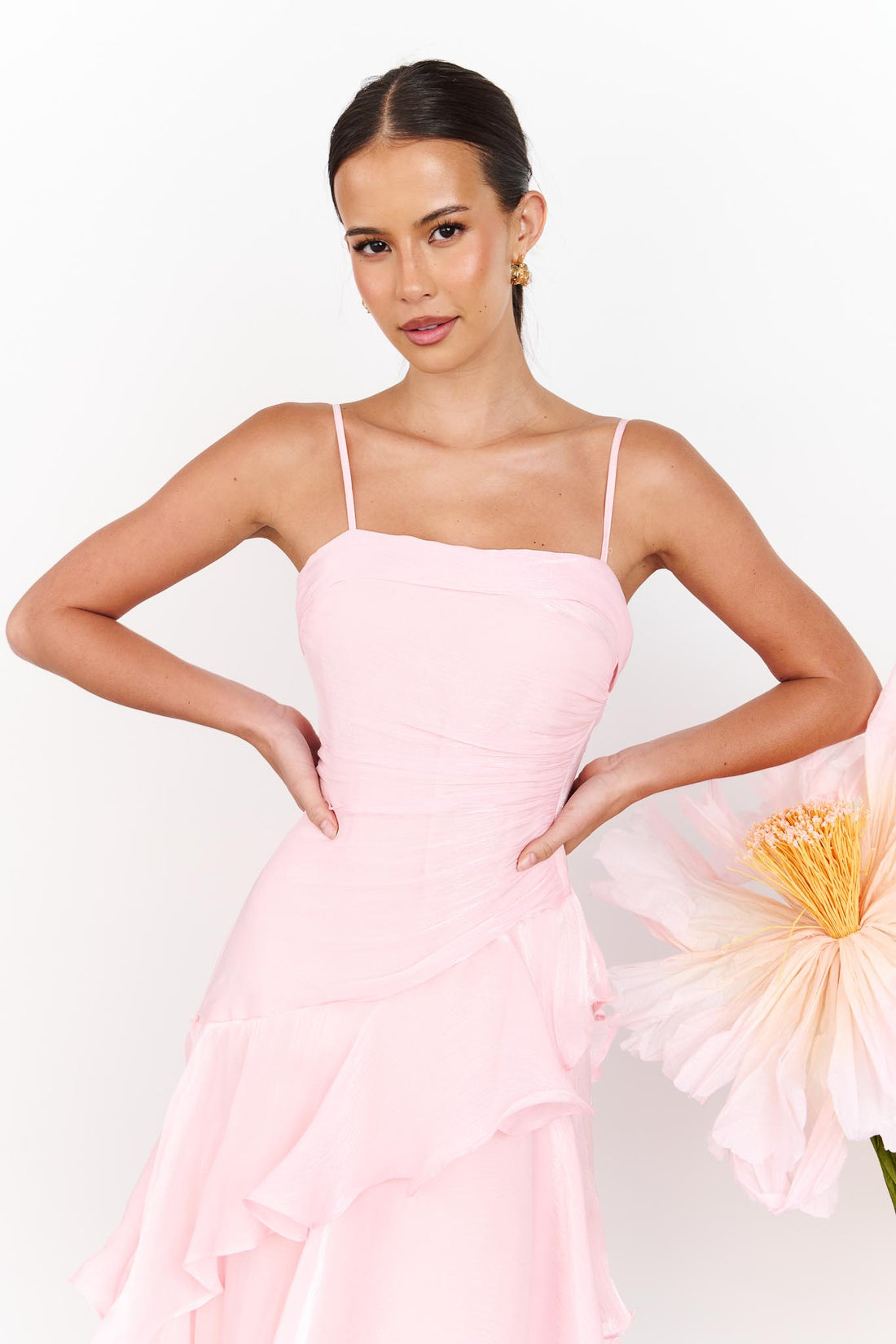 Close up of a woman wearing the minnie + MAXXIE Monaco maxi dress in light pink with a ruched bodice and layered ruffle skirt, standing against a white wall with a large pale pink flower on the right.