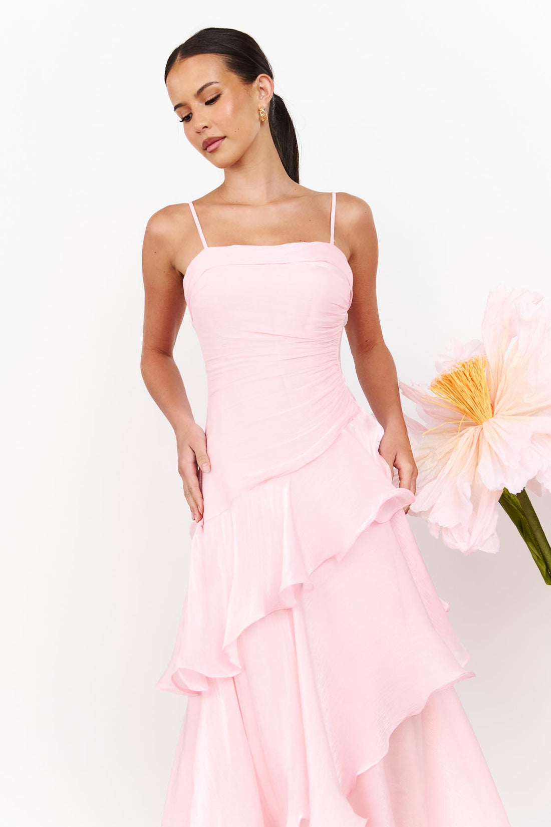 A woman wearing the minnie + MAXXIE Monaco maxi dress in light pink with a ruched bodice and layered ruffle skirt, standing against a white wall with a large pale pink flower on the right.