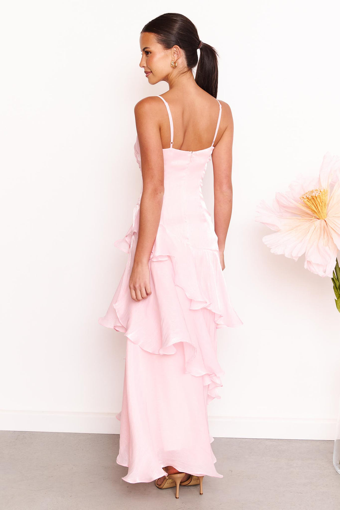 Back view of a woman wearing the minnie + MAXXIE Monaco maxi dress in light pink with a ruched bodice and layered ruffle skirt, standing against a white wall with a large pale pink flower on the right.