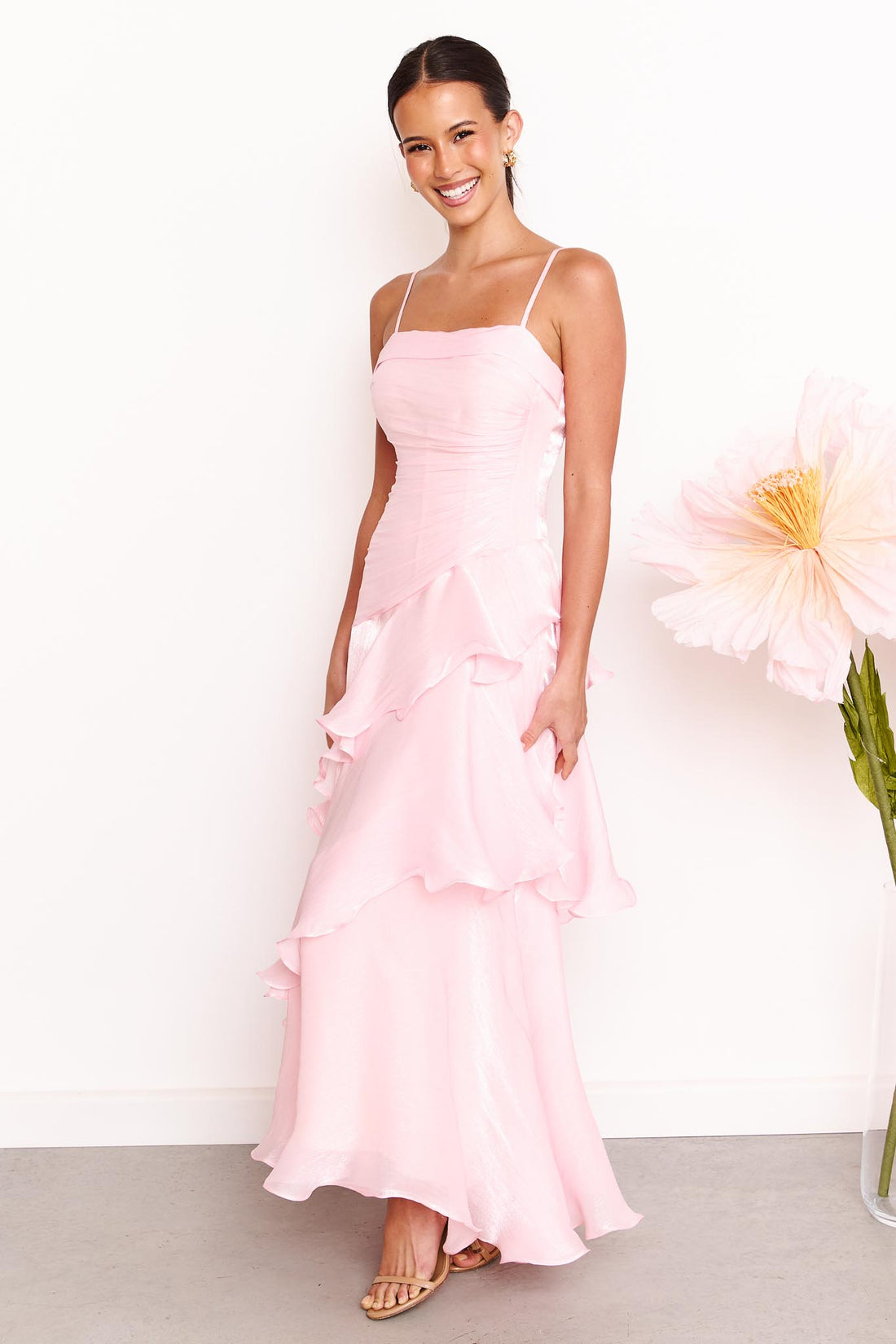 Side view of a woman wearing the minnie + MAXXIE Monaco maxi dress in light pink with a ruched bodice and layered ruffle skirt, standing against a white wall with a large pale pink flower on the right.