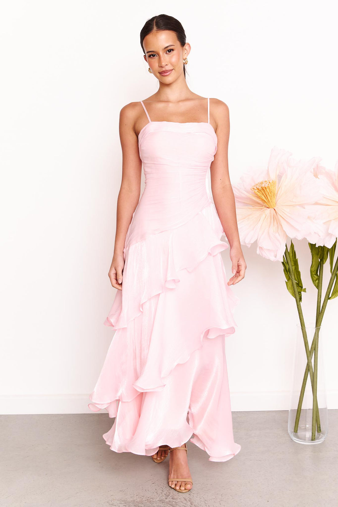 A woman wearing the minnie + MAXXIE Monaco maxi dress in light pink with a ruched bodice and layered ruffle skirt, standing against a white wall with a large pale pink flower on the right.