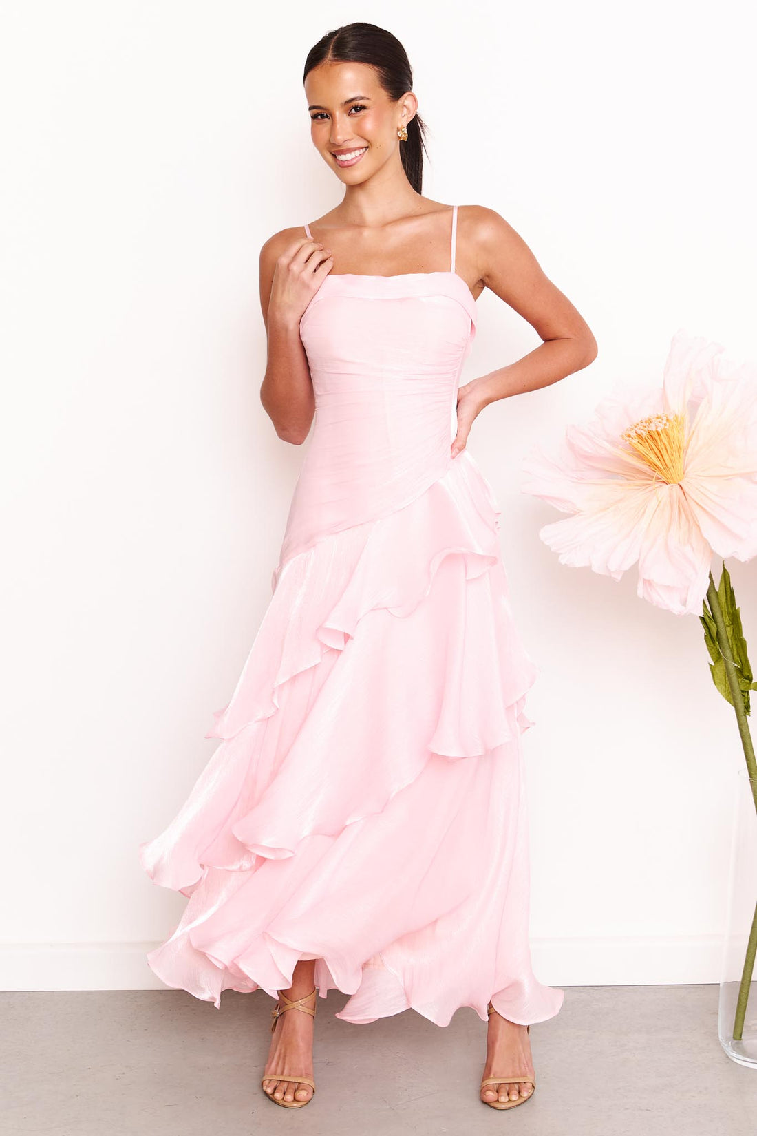 A woman wearing the minnie + MAXXIE Monaco maxi dress in light pink with a ruched bodice and layered ruffle skirt, standing against a white wall with a large pale pink flower on the right.