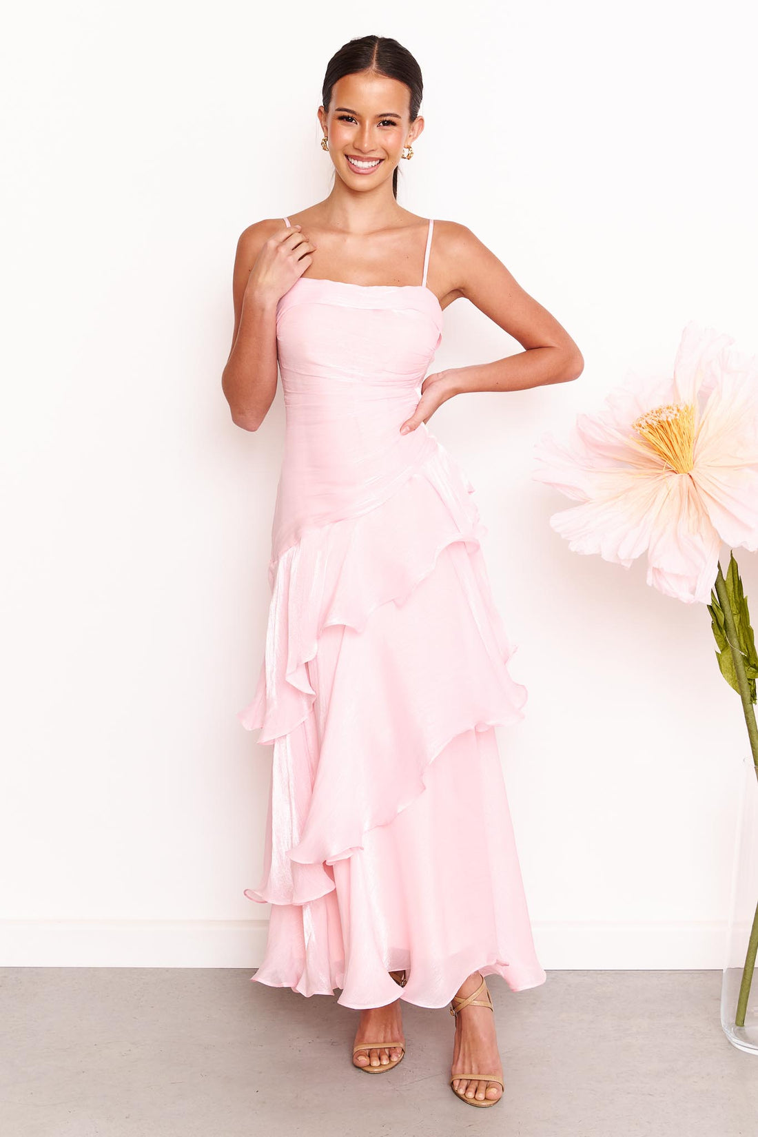 A woman wearing the minnie + MAXXIE Monaco maxi dress in light pink with a ruched bodice and layered ruffle skirt, standing against a white wall with a large pale pink flower on the right.