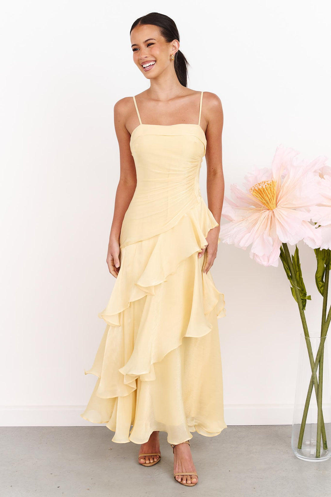 A woman wearing the minnie + MAXXIE Monaco Maxi Dress in pale yellow with a ruched bodice and layered ruffle skirt, standing against a white wall next to a vase of large pink flowers.