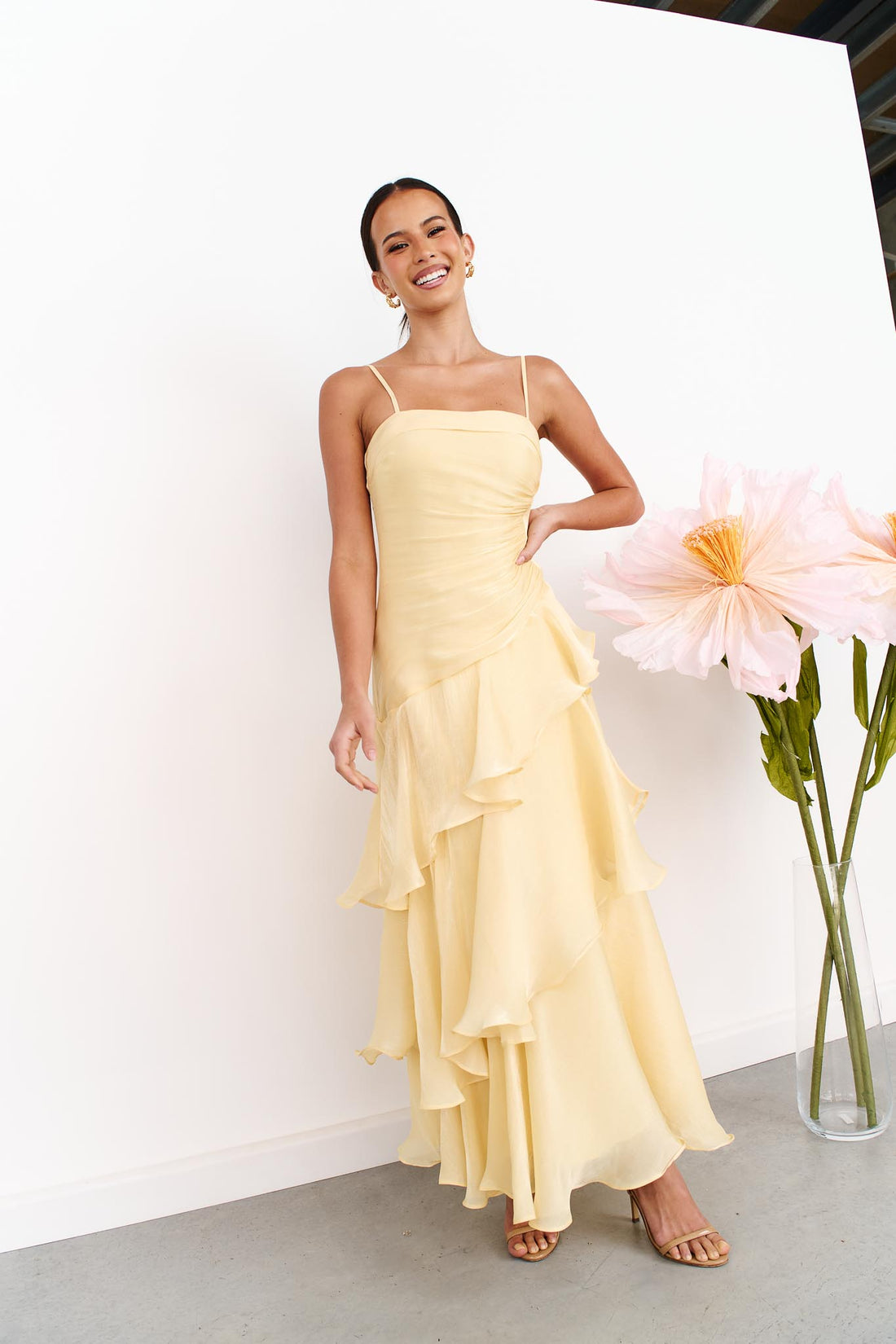 A woman wearing the minnie + MAXXIE Monaco Maxi Dress in pale yellow with a ruched bodice and layered ruffle skirt, standing against a white wall next to a vase of large pink flowers.