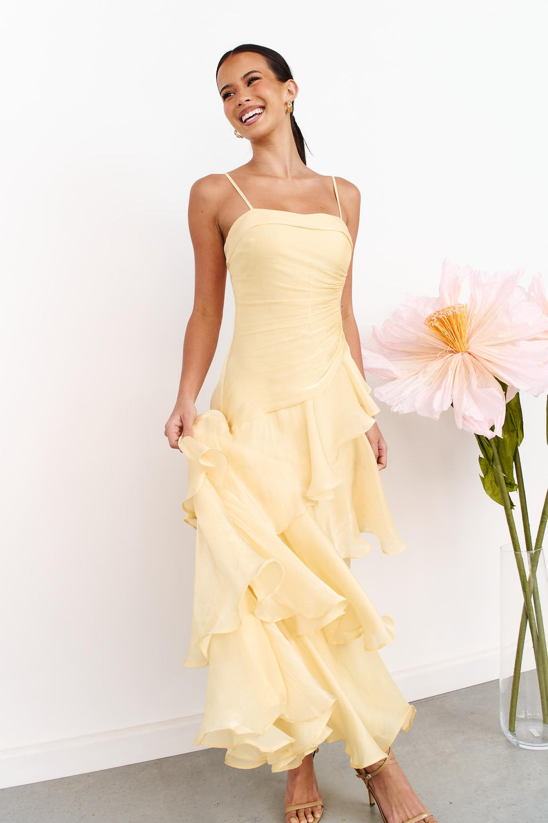 A woman wearing the minnie + MAXXIE Monaco Maxi Dress in pale yellow with a ruched bodice and layered ruffle skirt, standing against a white wall next to a vase of large pink flowers.