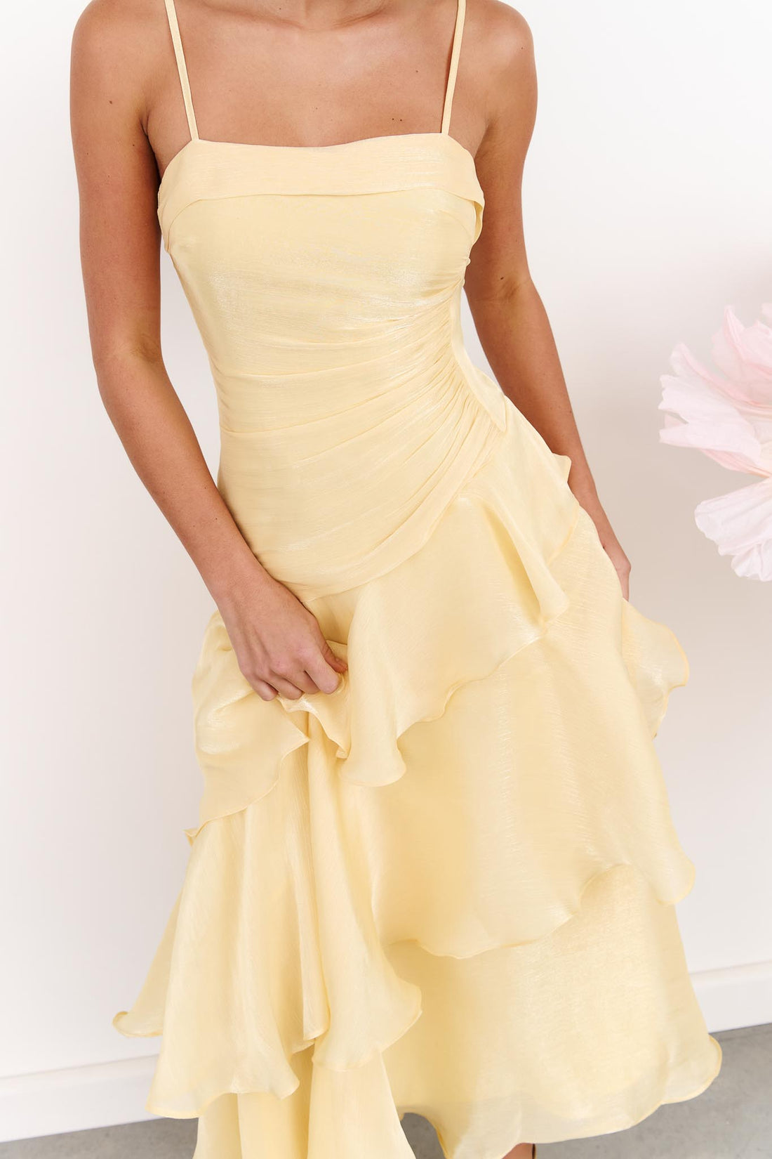 Detail of a woman wearing the minnie + MAXXIE Monaco Maxi Dress in pale yellow with a ruched bodice and layered ruffle skirt, standing against a white wall.