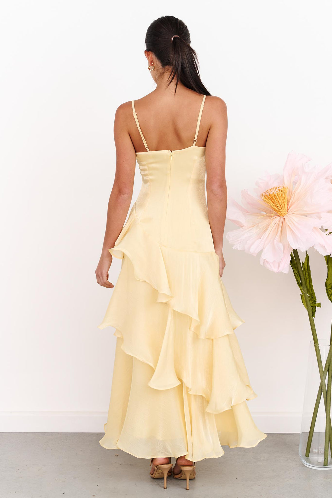 Back view of a woman wearing the minnie + MAXXIE Monaco Maxi Dress in pale yellow with a ruched bodice and layered ruffle skirt, standing against a white wall next to a vase of large pink flowers.