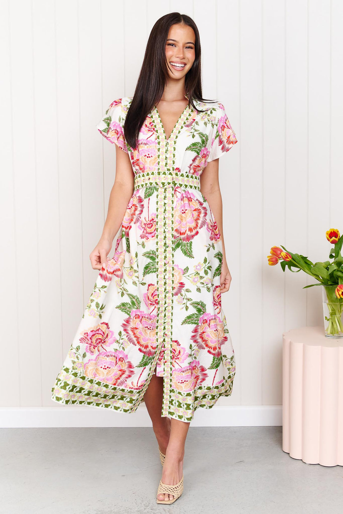 Woman wearing the minnie + MAXXIE Monet Midi Dress in a pink and green floral print with matching waist tie, standing against a white wall.