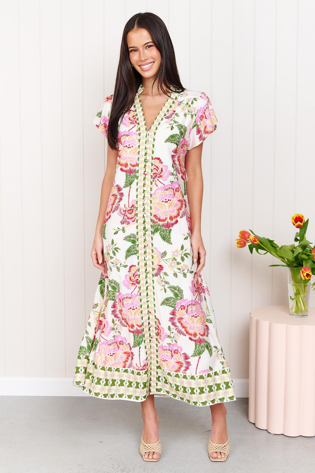 Woman wearing the minnie + MAXXIE Monet Midi Dress in a pink and green floral print, standing against a white wall.