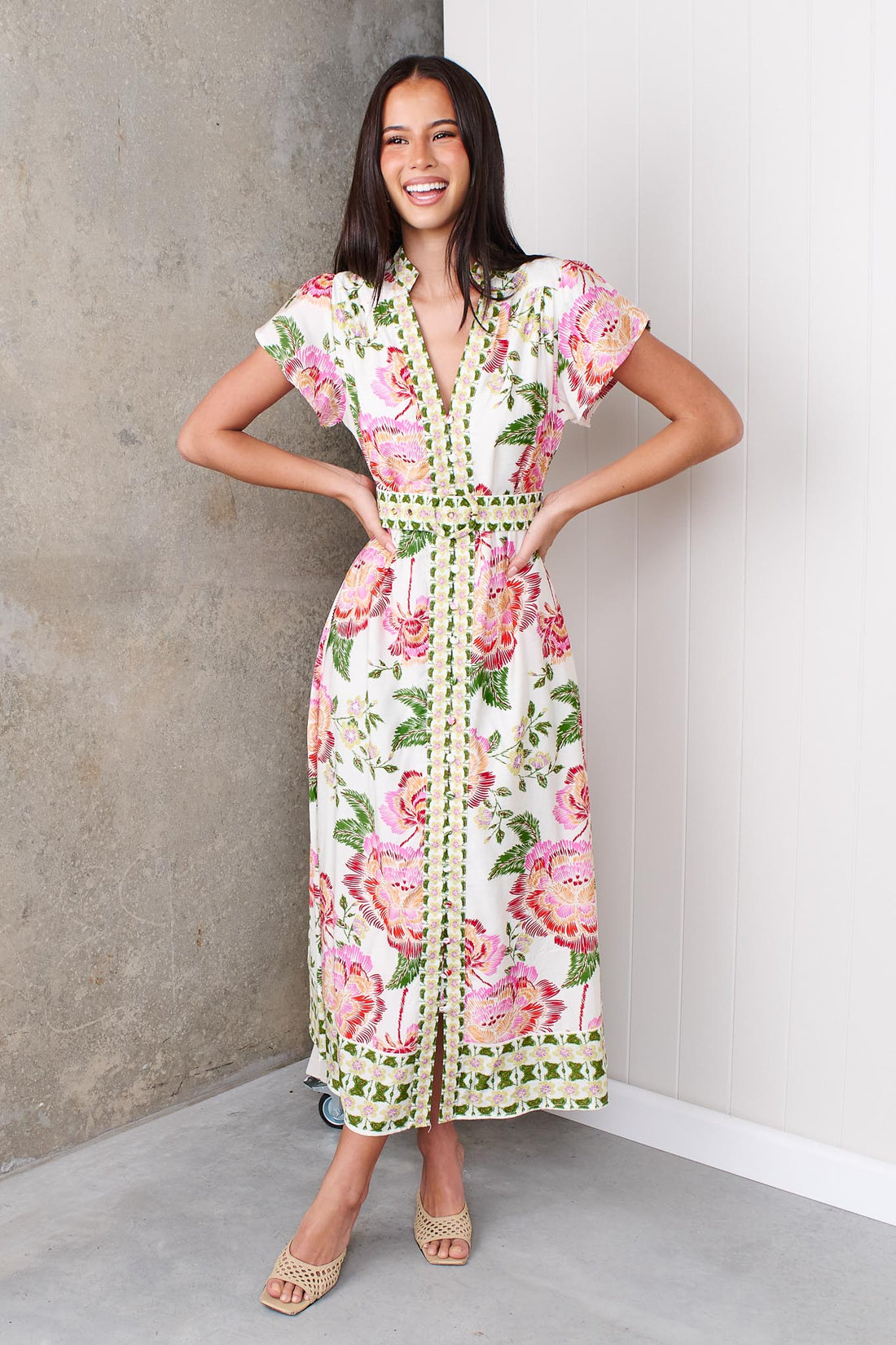 Woman wearing the minnie + MAXXIE Monet Midi Dress in a pink and green floral print with matching waist tie, standing against a white and concrete wall.