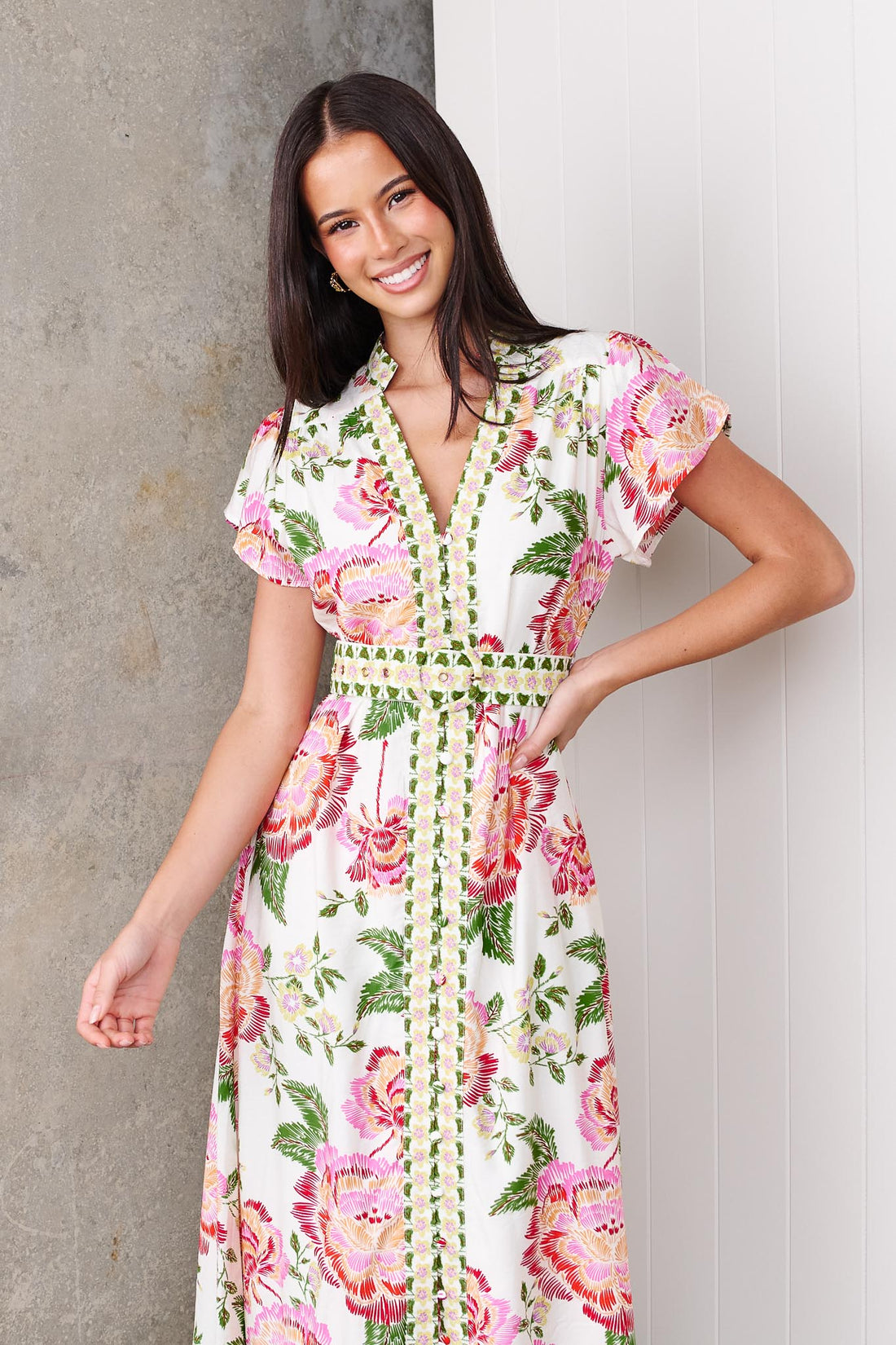 Woman wearing the minnie + MAXXIE Monet Midi Dress in a pink and green floral print with matching waist tie, standing against a white wall.