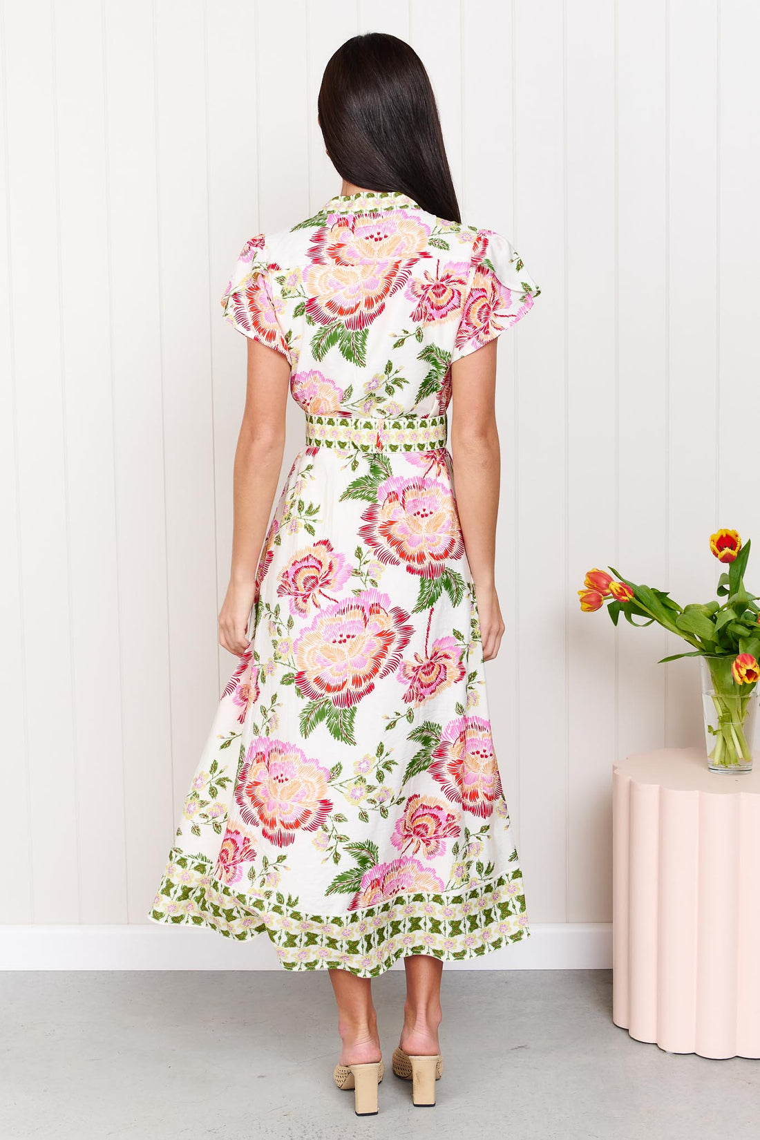 Back view of a woman wearing the minnie + MAXXIE Monet Midi Dress in a pink and green floral print with matching waist tie, standing against a white wall.