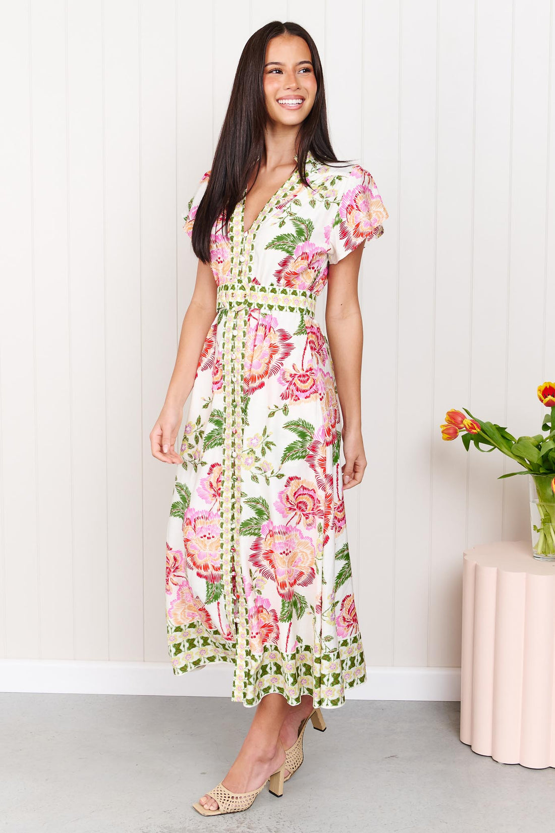 Woman wearing the minnie + MAXXIE Monet Midi Dress in a pink and green floral print with matching waist tie, standing against a white wall.