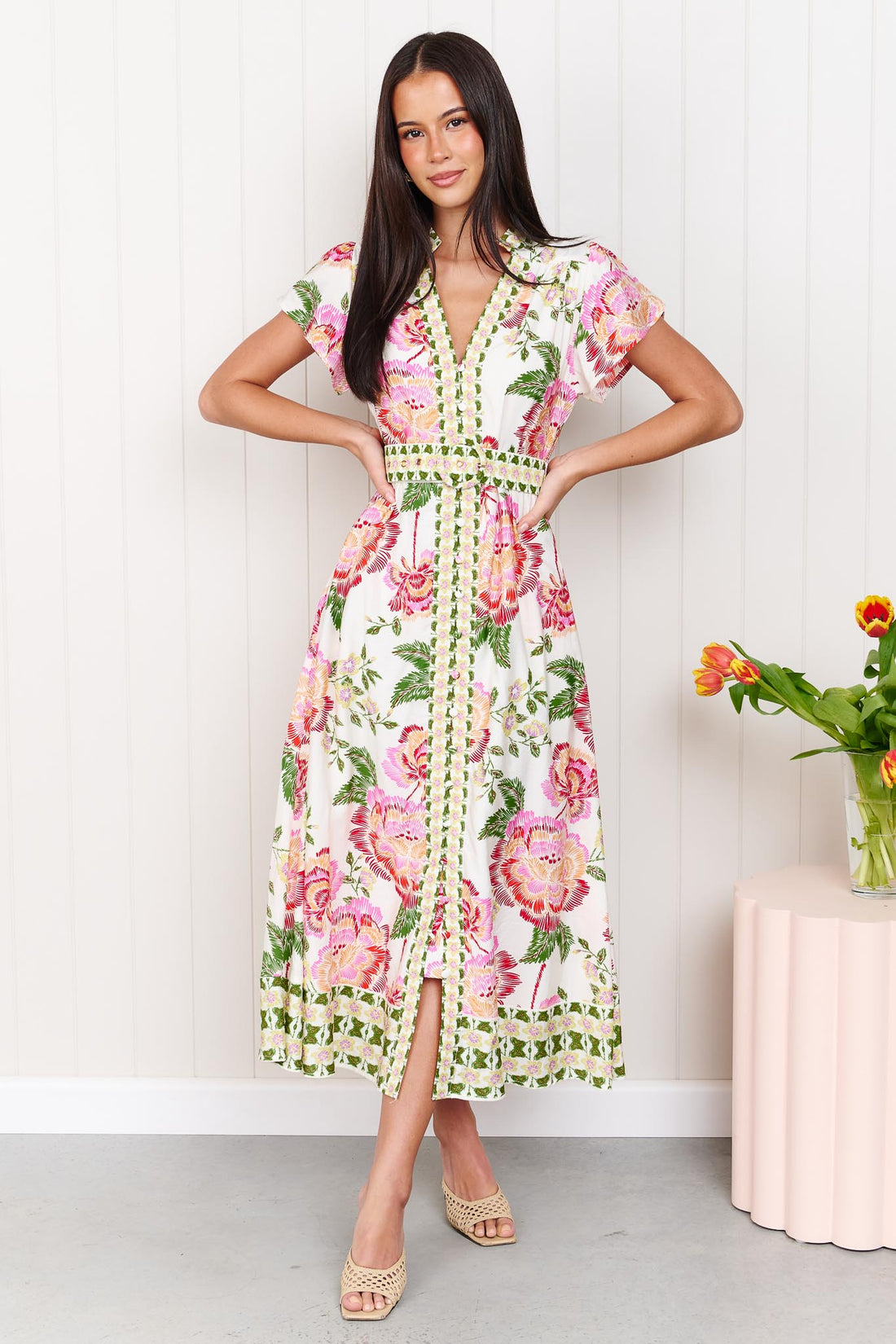 Woman wearing the minnie + MAXXIE Monet Midi Dress in a pink and green floral print with matching waist tie, standing against a white wall.