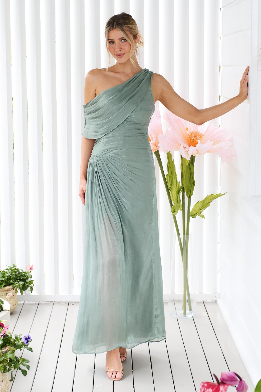 A woman wearing the minnie + MAXXIE Opera off-shoulder maxi event dress in sage green, standing against a white panelled wall with flowers.