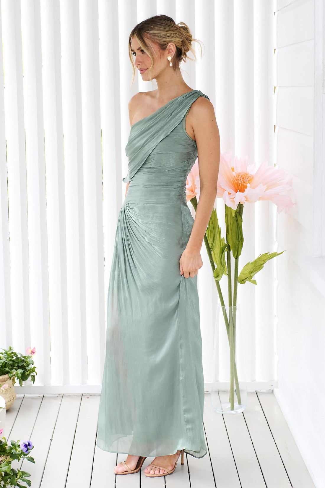 Side view of a woman wearing the minnie + MAXXIE Opera off-shoulder maxi event dress in sage green, standing against a white panelled wall with flowers.