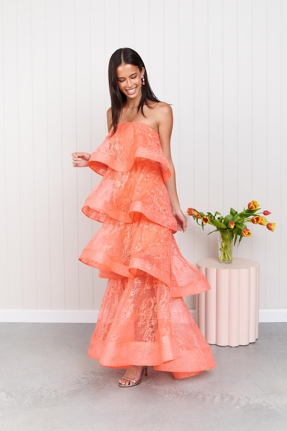 A woman wearing the Pasduchas Ophelia Lace tiered maxi event dress in sunkissed coral orange lace, standing in front of a white panelled wall.