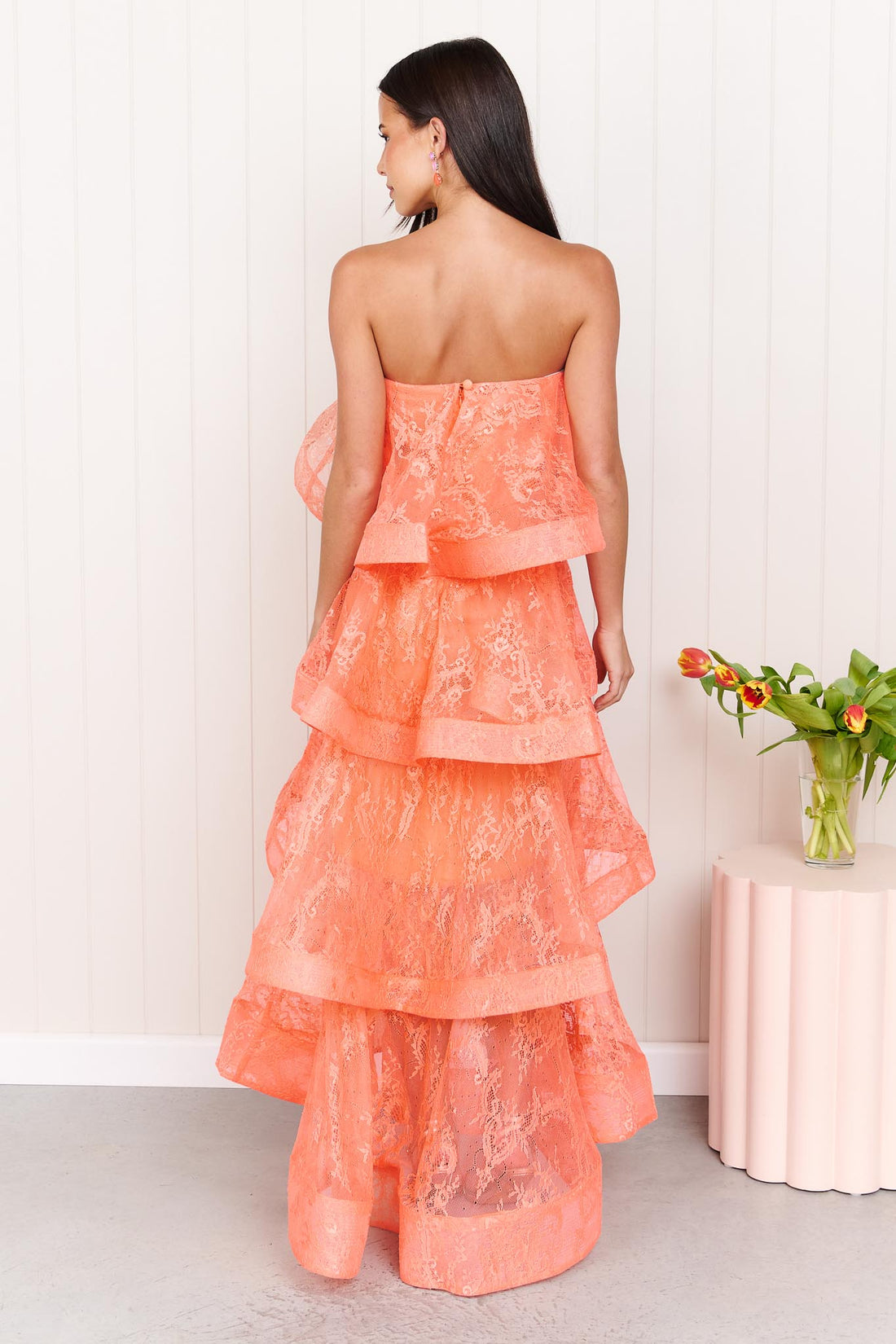 Back view of a woman wearing the Pasduchas Ophelia Lace tiered maxi event dress in sunkissed coral orange lace, standing in front of a white panelled wall.