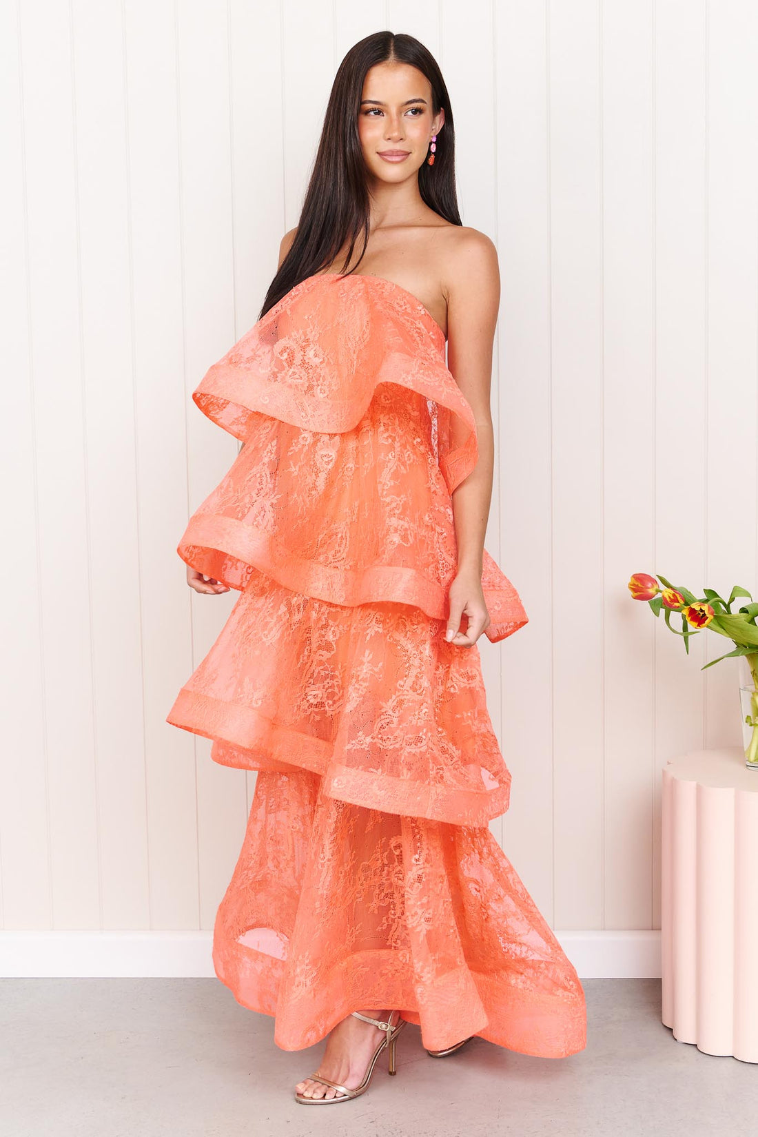 A woman wearing the Pasduchas Ophelia Lace tiered maxi event dress in sunkissed coral orange lace, standing in front of a white panelled wall.