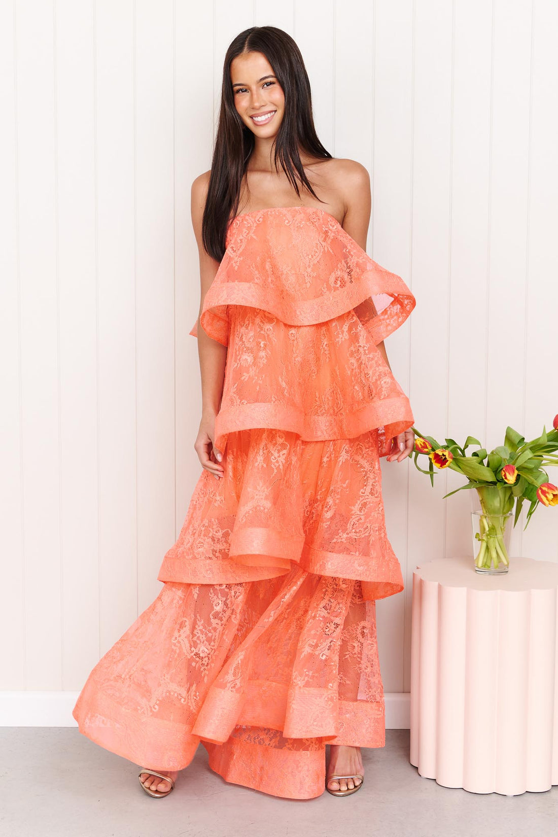 A woman wearing the Pasduchas Ophelia Lace tiered maxi event dress in sunkissed coral orange lace, standing in front of a white panelled wall.