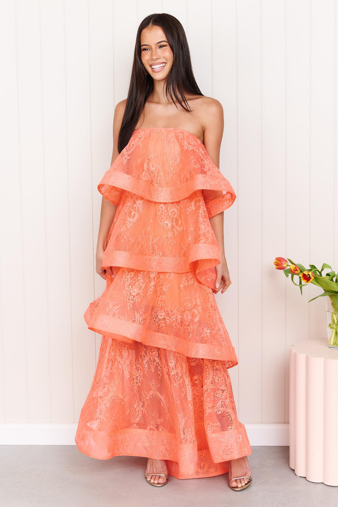 A woman wearing the Pasduchas Ophelia Lace tiered maxi event dress in sunkissed coral orange lace, standing in front of a white panelled wall.
