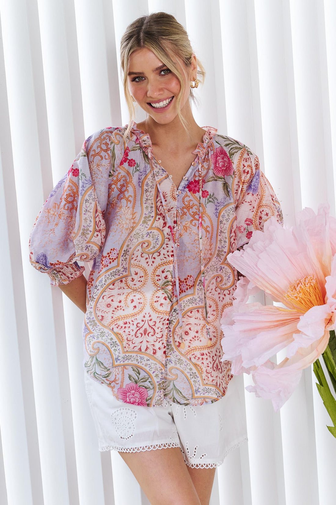 A woman wearing the minnie + MAXXIE Paloma paisley printed blouse in pinks and lilacs, standing against a white panelled wall.
