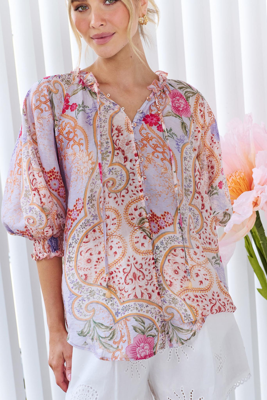 A woman wearing the minnie + MAXXIE Paloma paisley printed blouse in pinks and lilacs, standing against a white panelled wall.