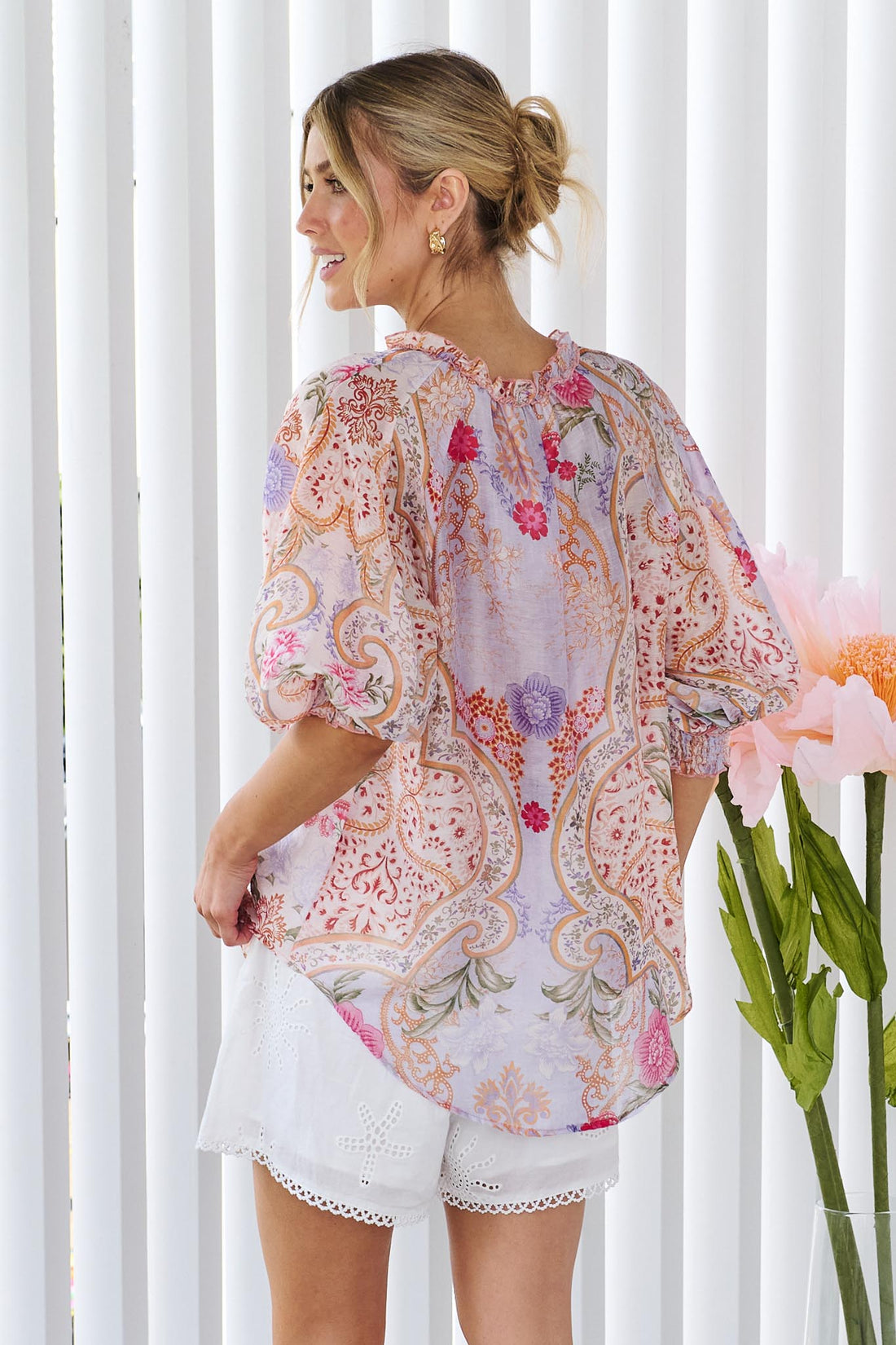 Back view of a woman wearing the minnie + MAXXIE Paloma paisley printed blouse in pinks and lilacs, standing against a white panelled wall.
