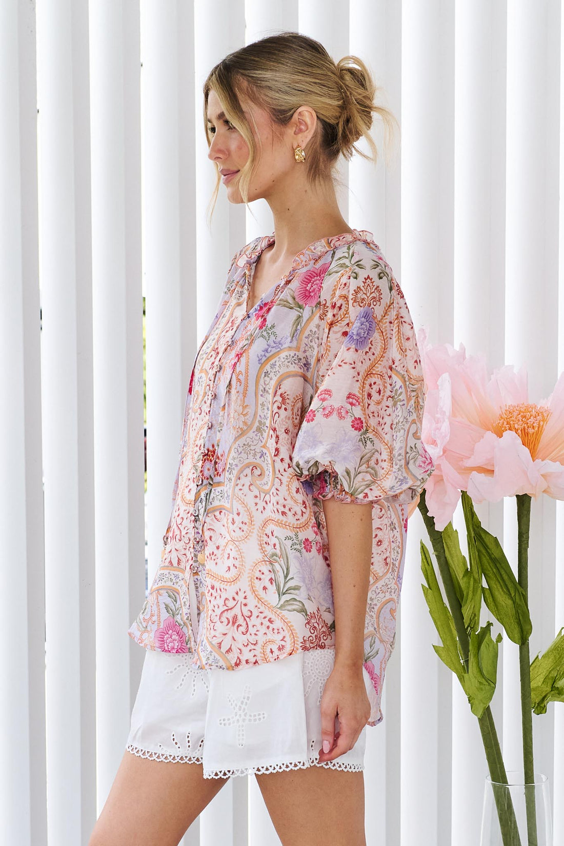 Side view of a woman wearing the minnie + MAXXIE Paloma paisley printed blouse in pinks and lilacs, standing against a white panelled wall.