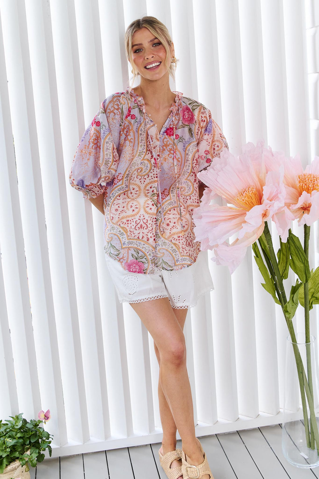 A woman wearing the minnie + MAXXIE Paloma paisley printed blouse in pinks and lilacs, standing against a white panelled wall.