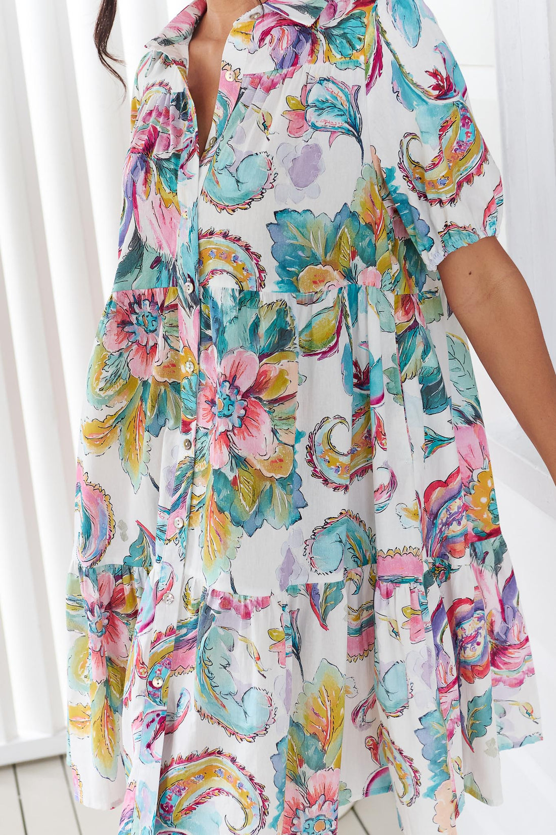 Detail of a woman wearing the minnie + MAXXIE Penny short-sleeve a-line dress with a colourful watercolour floral and paisley print, standing in front of a white panelled wall.