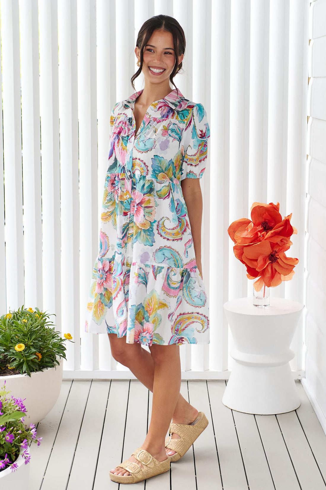 A woman wearing the minnie + MAXXIE Penny short-sleeve a-line dress with a colourful watercolour floral and paisley print, standing in front of a white panelled wall.