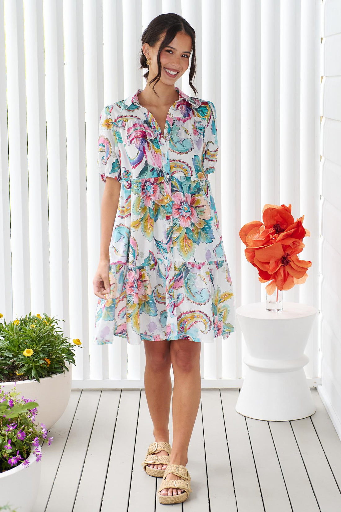 A woman wearing the minnie + MAXXIE Penny short-sleeve a-line dress with a colourful watercolour floral and paisley print, standing in front of a white panelled wall.