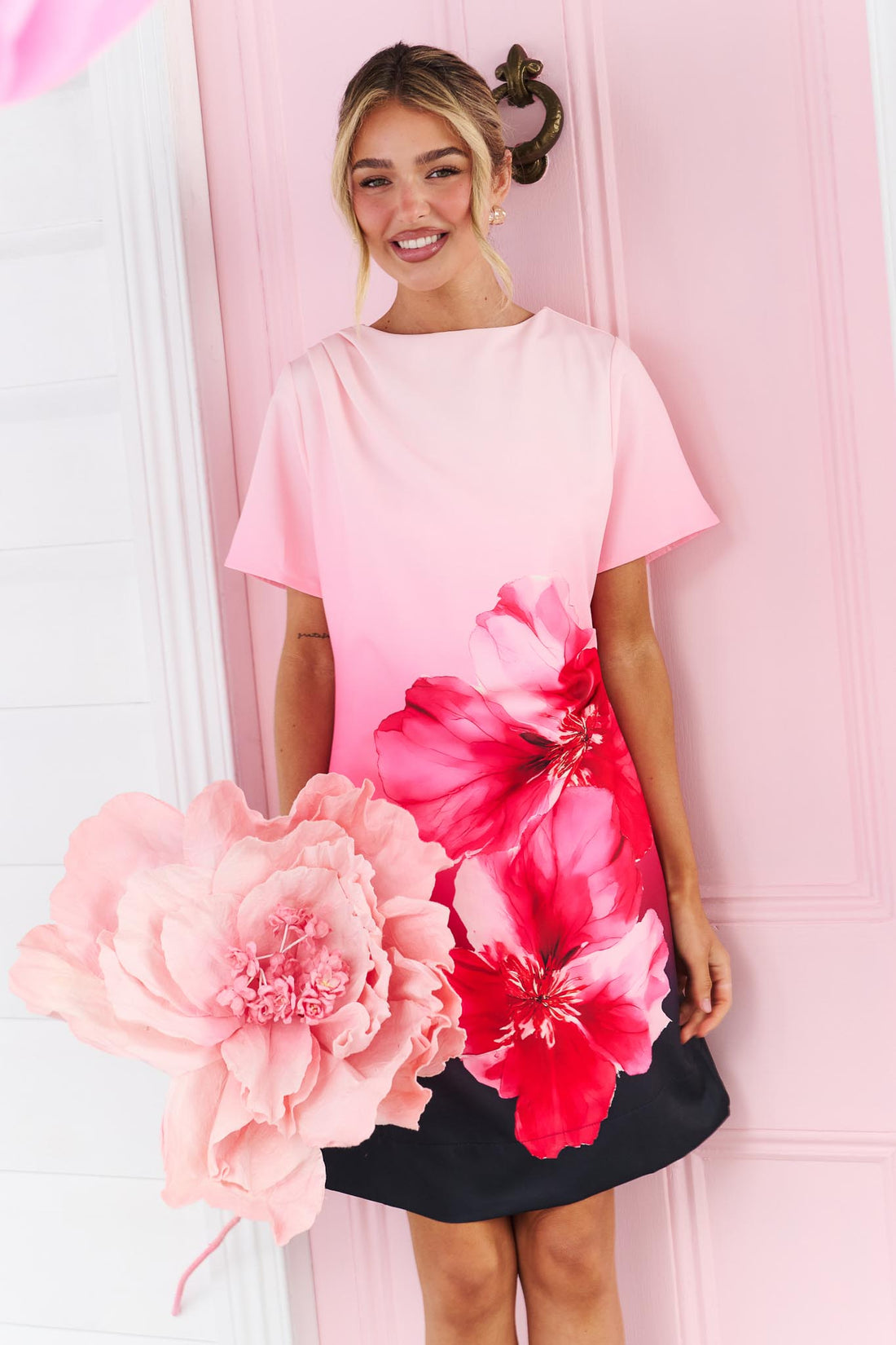 A woman wearing the minnie + MAXXIE short-sleeve A-line Peony dress with a pink-to-black ombre gradient and a large red hibiscus flower print on the front, standing in front of a pink door.
