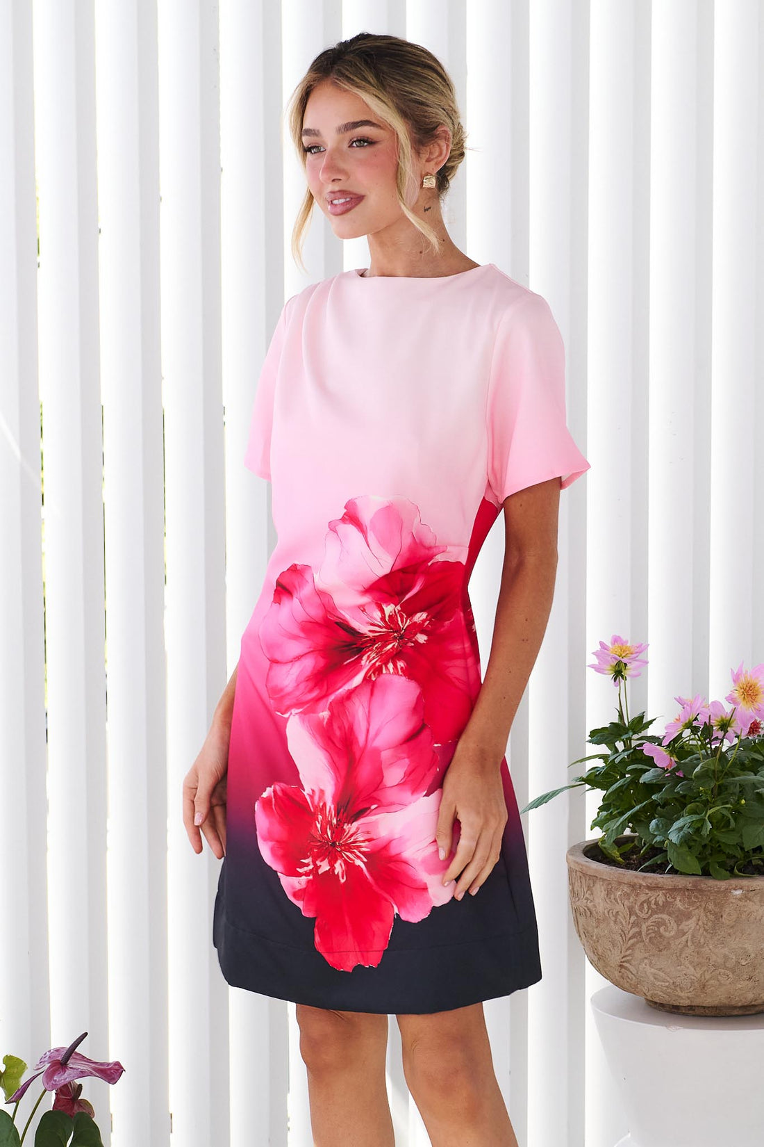 Side view of a woman wearing the minnie + MAXXIE short-sleeve A-line Peony dress with a pink-to-black ombre gradient and a large red hibiscus flower print on the front, standing in front of a white panel wall.
