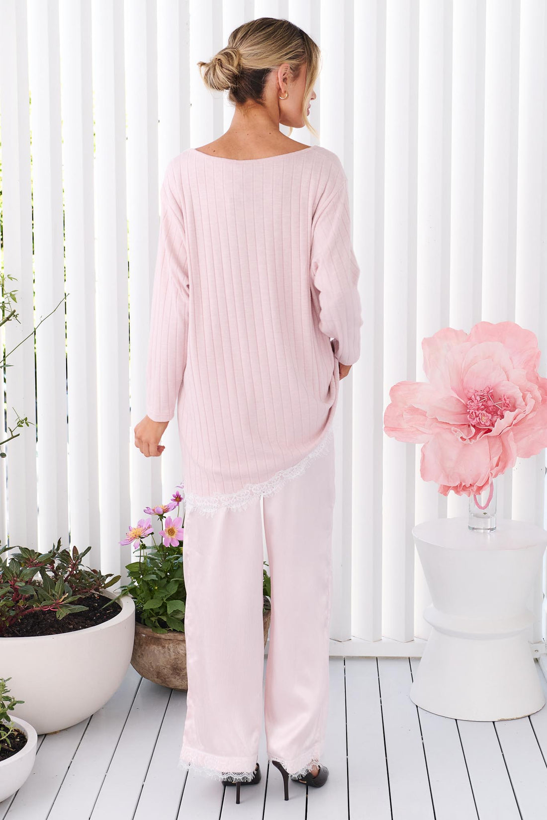 Back view of a woman wearing the minnie + MAXXIE pink Lila Lace Trim Satin Pant and pink long-sleeve top, standing in front of a white slatted wall with decorative plants and a large pink flower prop.