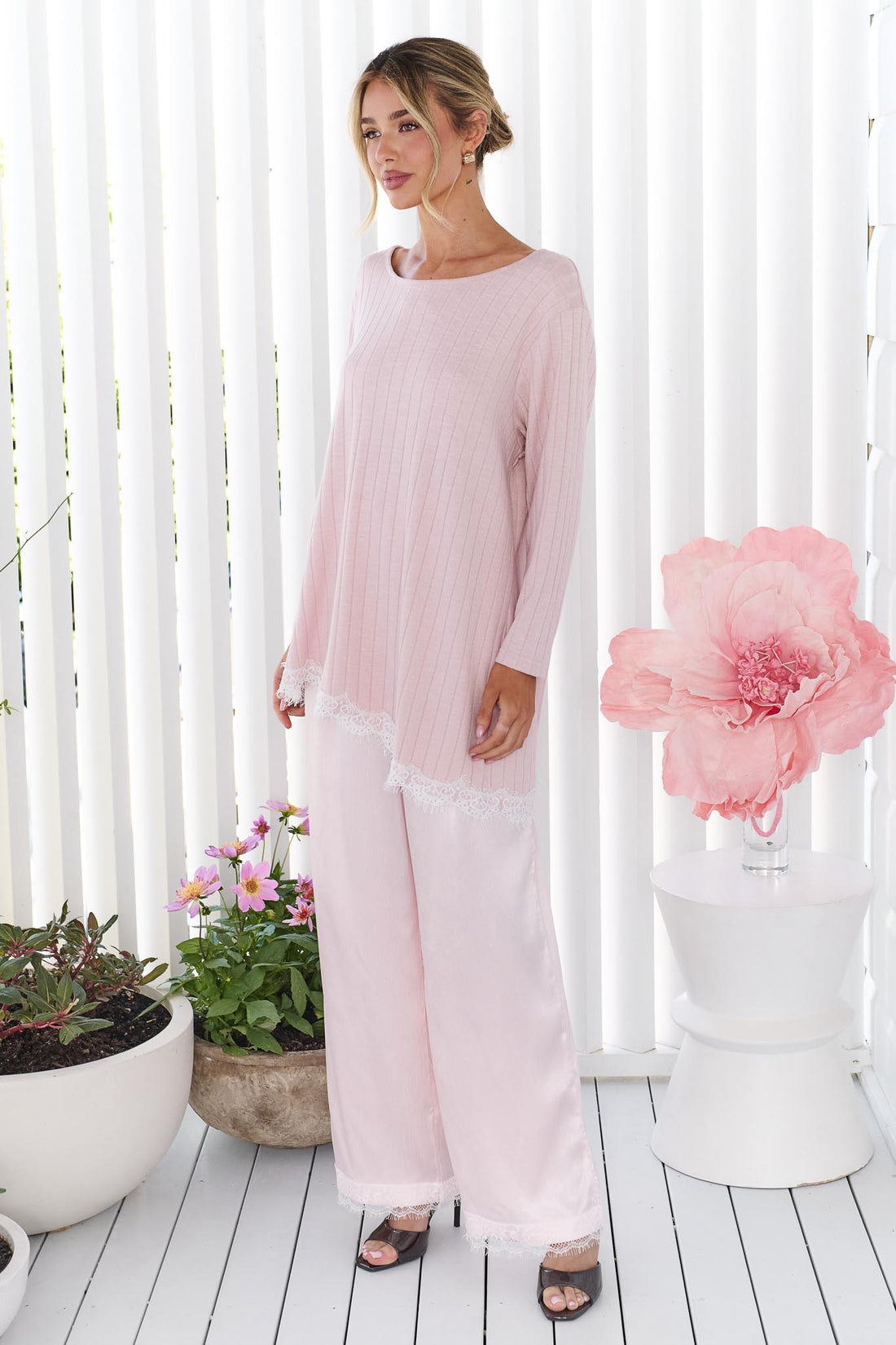 A woman wearing the minnie + MAXXIE pink Lila Lace Trim Satin Pant and pink long-sleeve top, standing in front of a white slatted wall with decorative plants and a large pink flower prop.