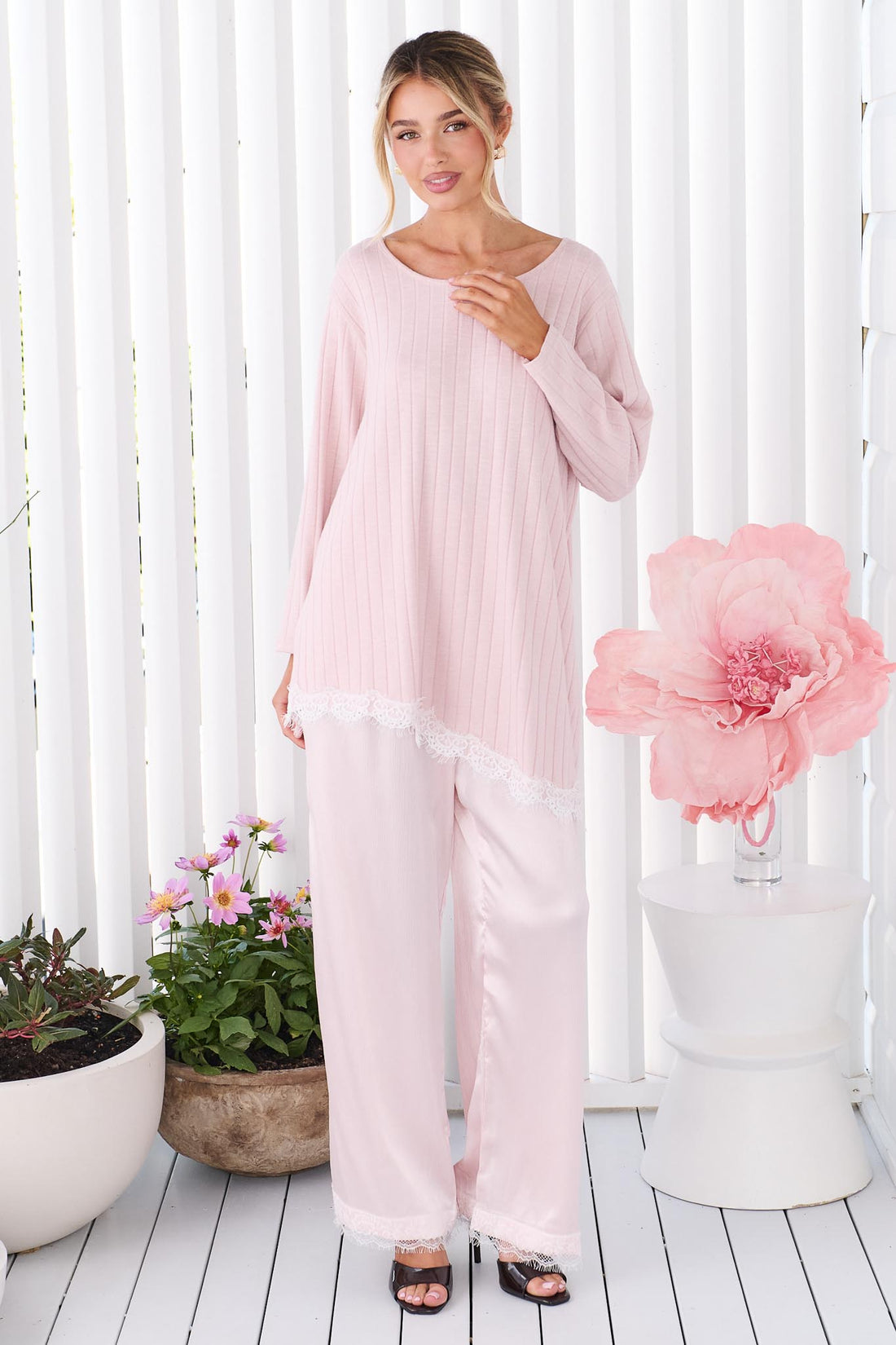 A woman wearing the minnie + MAXXIE pink Lila Lace Trim Satin Pant and pink long-sleeve top, standing in front of a white slatted wall with decorative plants and a large pink flower prop.