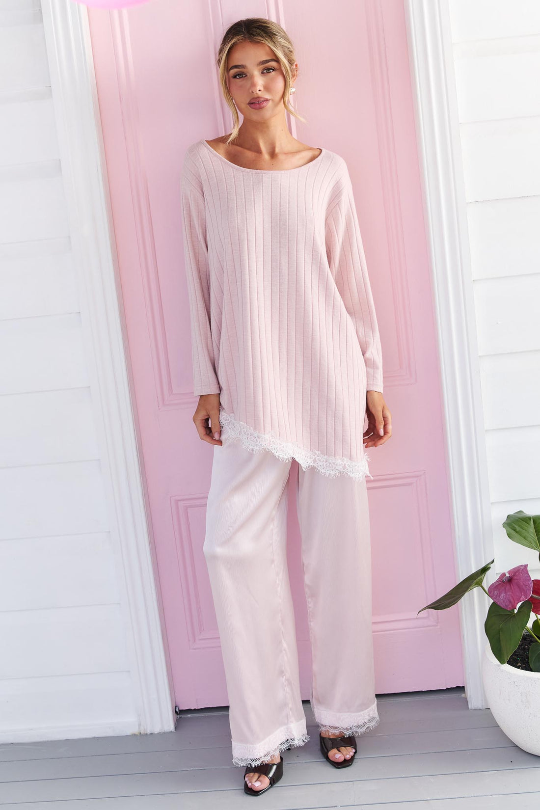 A woman wearing the minnie + MAXXIE pink Lila Lace Trim Satin Pant and pink long-sleeve top, standing in front of a pink door.