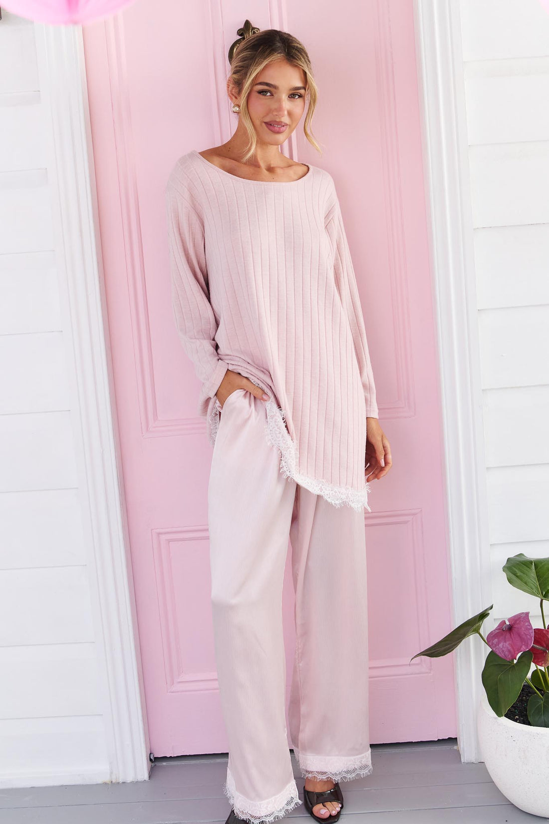 A woman wearing the minnie + MAXXIE pink Lila Lace Trim Satin Pant and pink long-sleeve top, standing in front of a pink door.