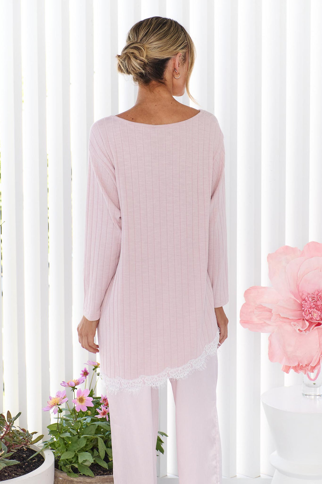 Back view of a woman wearing the minnie + MAXXIE Alina light pink ribbed long-sleeve tunic top with matching pants, standing in front of white vertical wall panels with decorative flowers nearby.