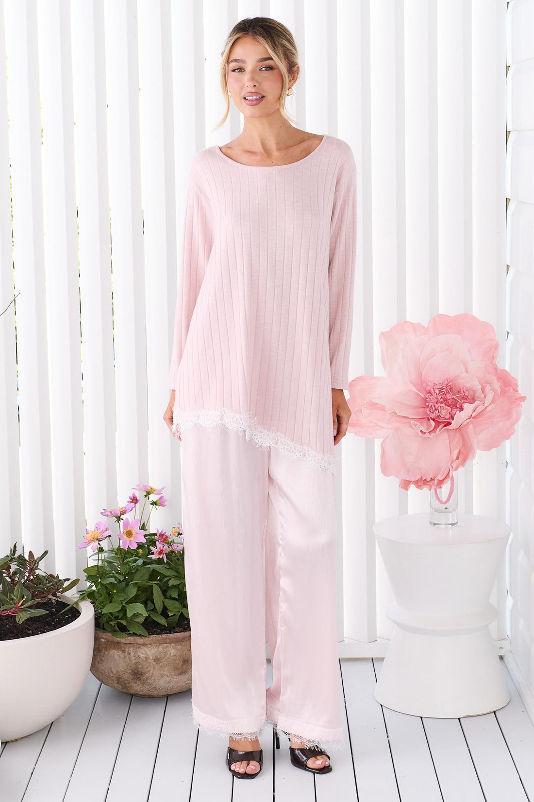 A woman wearing the minnie + MAXXIE Alina light pink long-sleeve tunic top with a round neckline and a lace hem, paired with matching pink satin pants, standing in front of white vertical panelled wall.