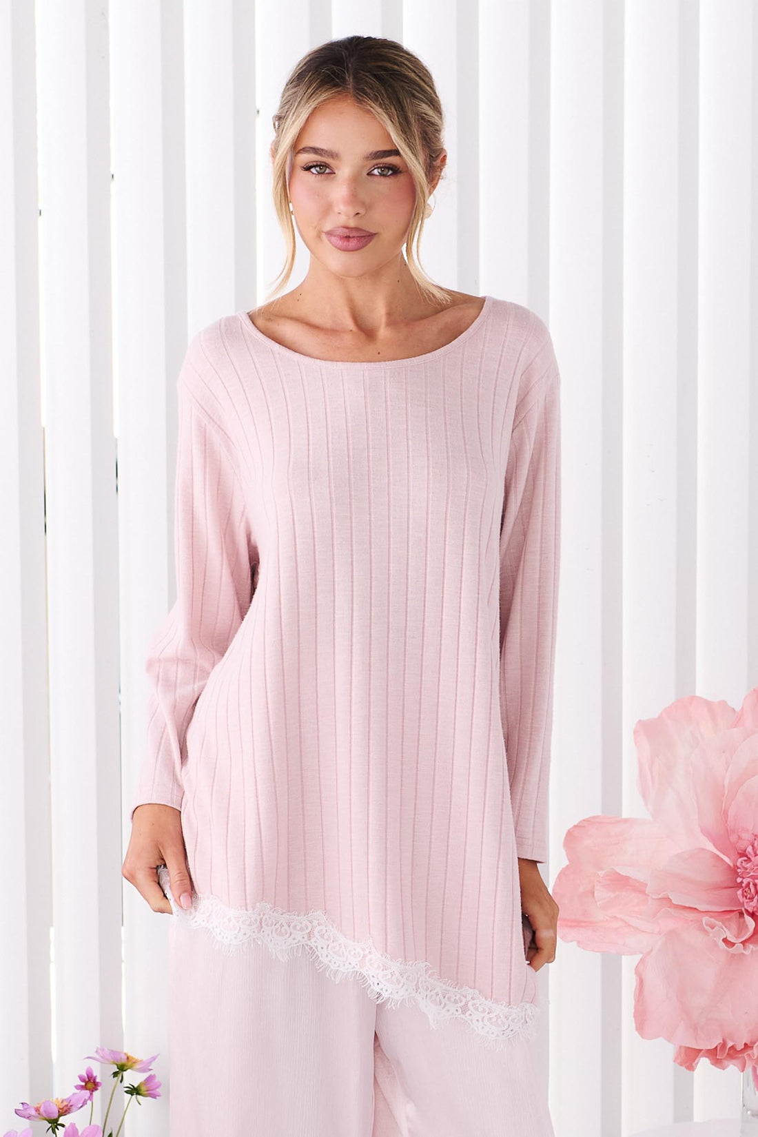 A woman wearing the minnie + MAXXIE Alina light pink long-sleeve tunic top with a round neckline and a lace hem, standing in front of white vertical panelled wall.