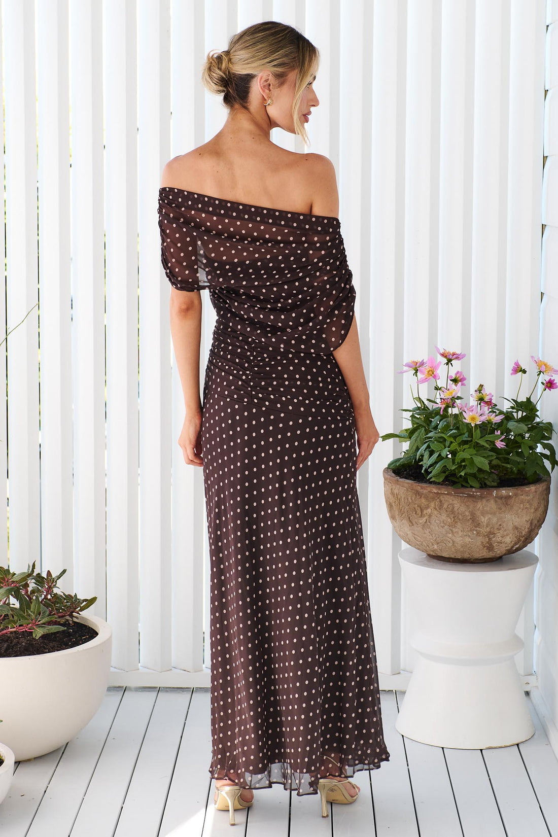Back view of a woman wearing the minnie + MAXXIE Pinto off-shoulder, maxi dark brown dress with polka dots, standing in front of a white vertical panelled wall.