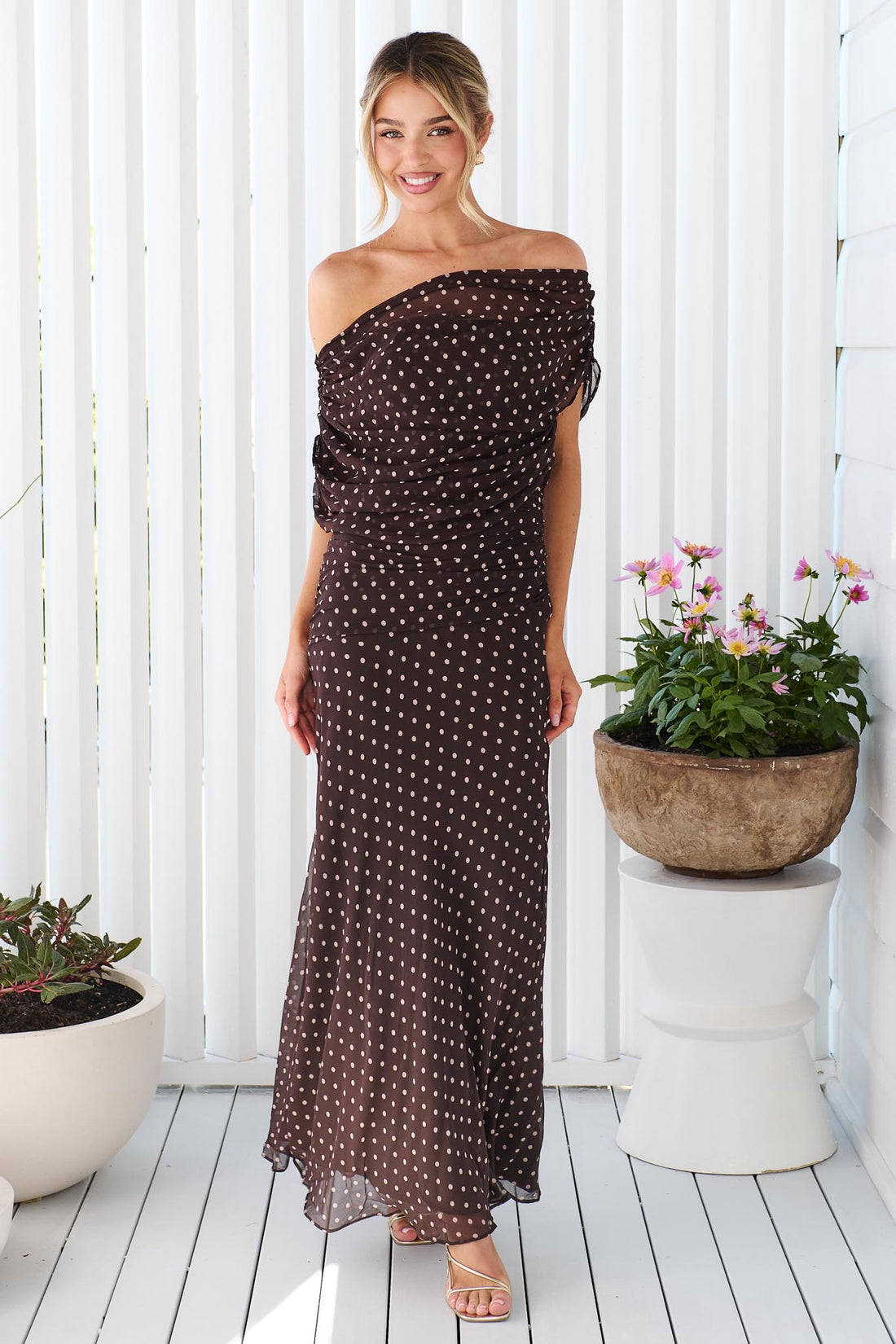 A woman wearing the minnie + MAXXIE Pinto off-shoulder, maxi dark brown dress with polka dots, standing in front of a white vertical panelled wall.